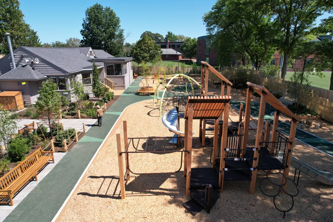 Playground with wooden structures, slides, and mulch ground cover, next to a building and trees.