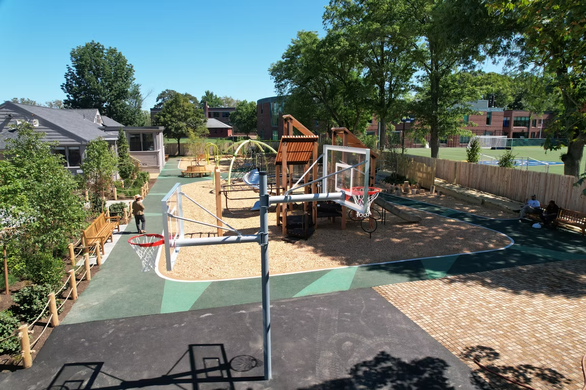 Playground with basketball court and wooden structures, trees, and buildings in the background.