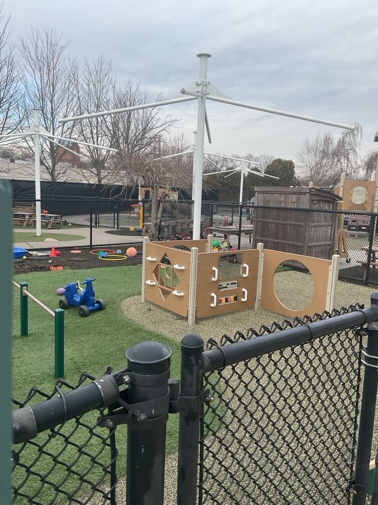 Playground with wooden structures, netting, and toys on green grass, behind a black chain-link fence.