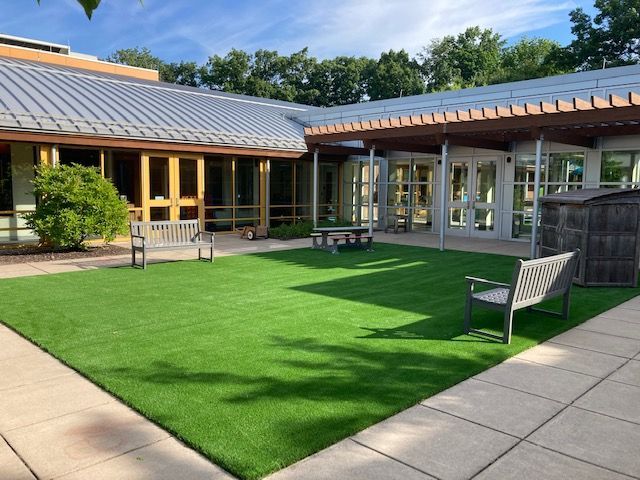 Courtyard with green turf, benches, and building with glass doors and a metal roof.