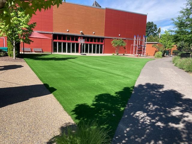 Green lawn with a red and orange building, a sidewalk, and trees.