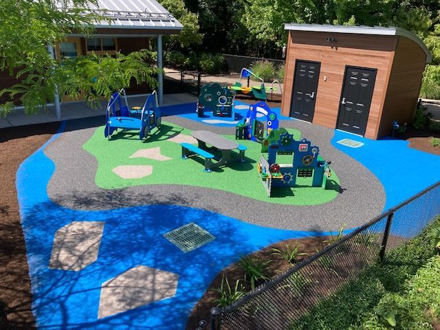 Playground with blue, green, and gray soft surfaces. Includes play structures and wooden building.