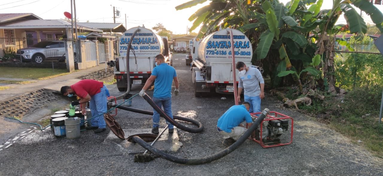 Hombres trabajando, limpiando el drenaje de aguas residuales con camiones, mangueras y bombas en una carretera.