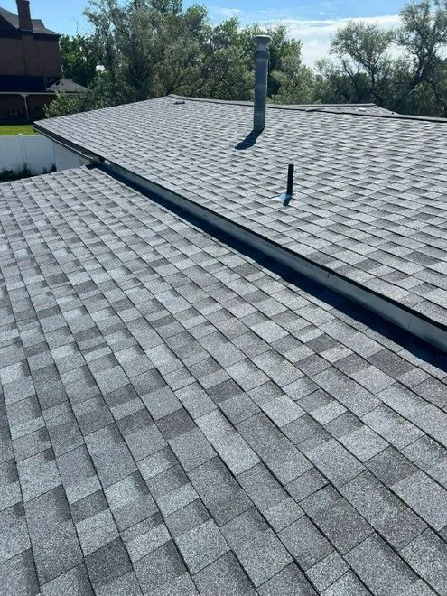 Gray asphalt shingle roof with a metal channel and two pipes; clear sky in the background.