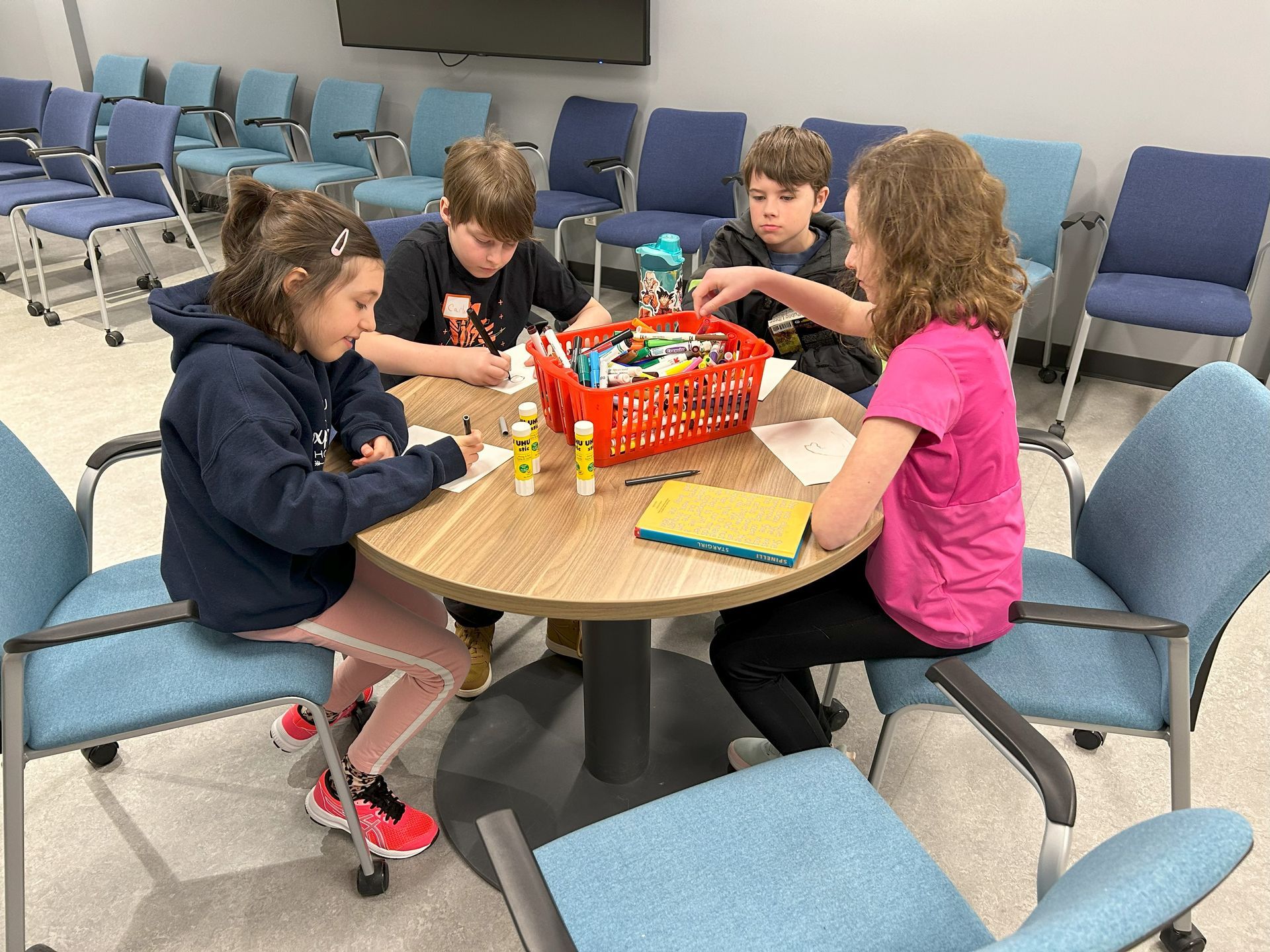 Four children sit at a round table, crafting with supplies. Blue chairs surround them in a room.