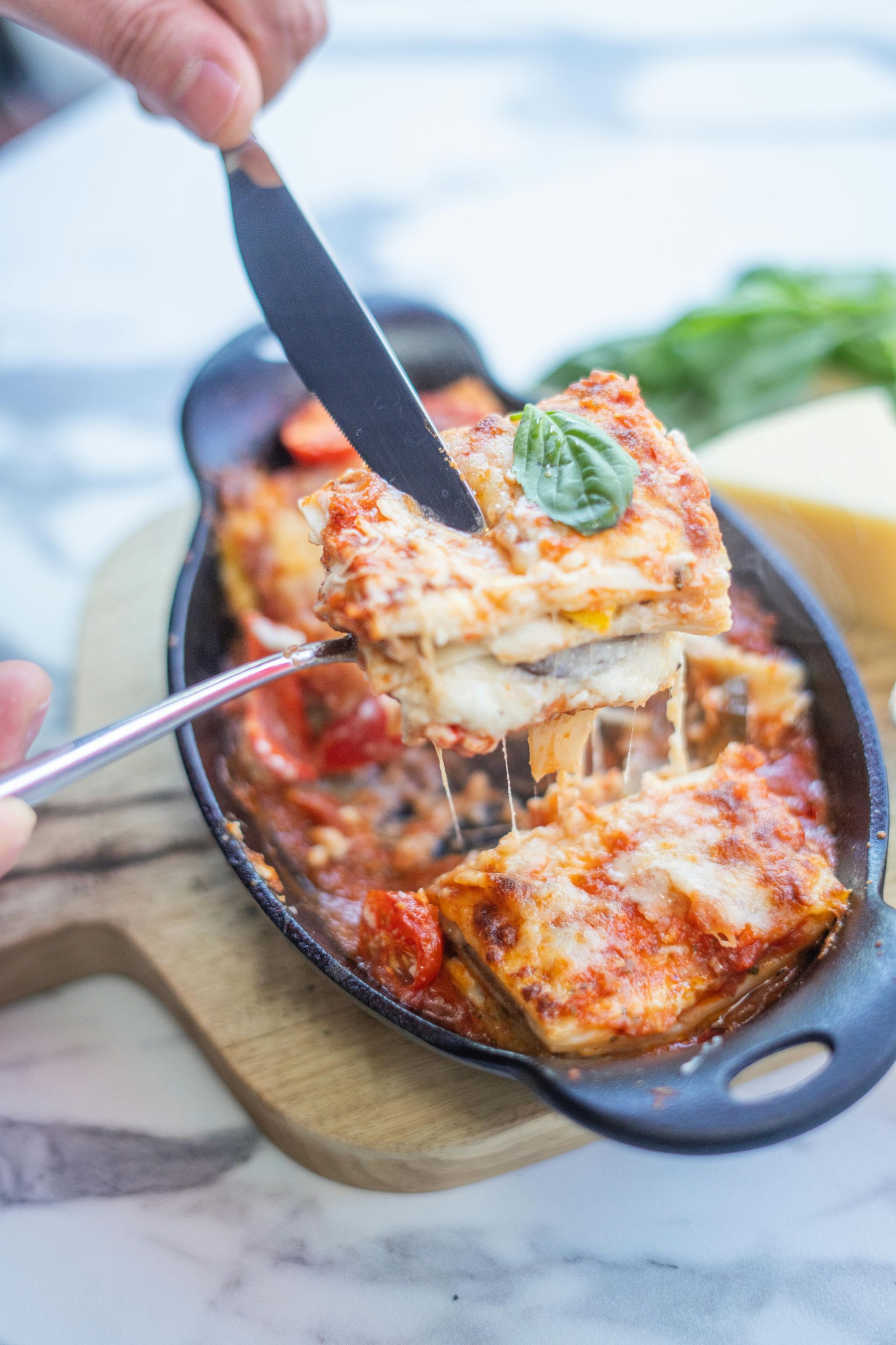 Lasagna being cut with a knife, cheesy and saucy in a black dish, garnished with basil.