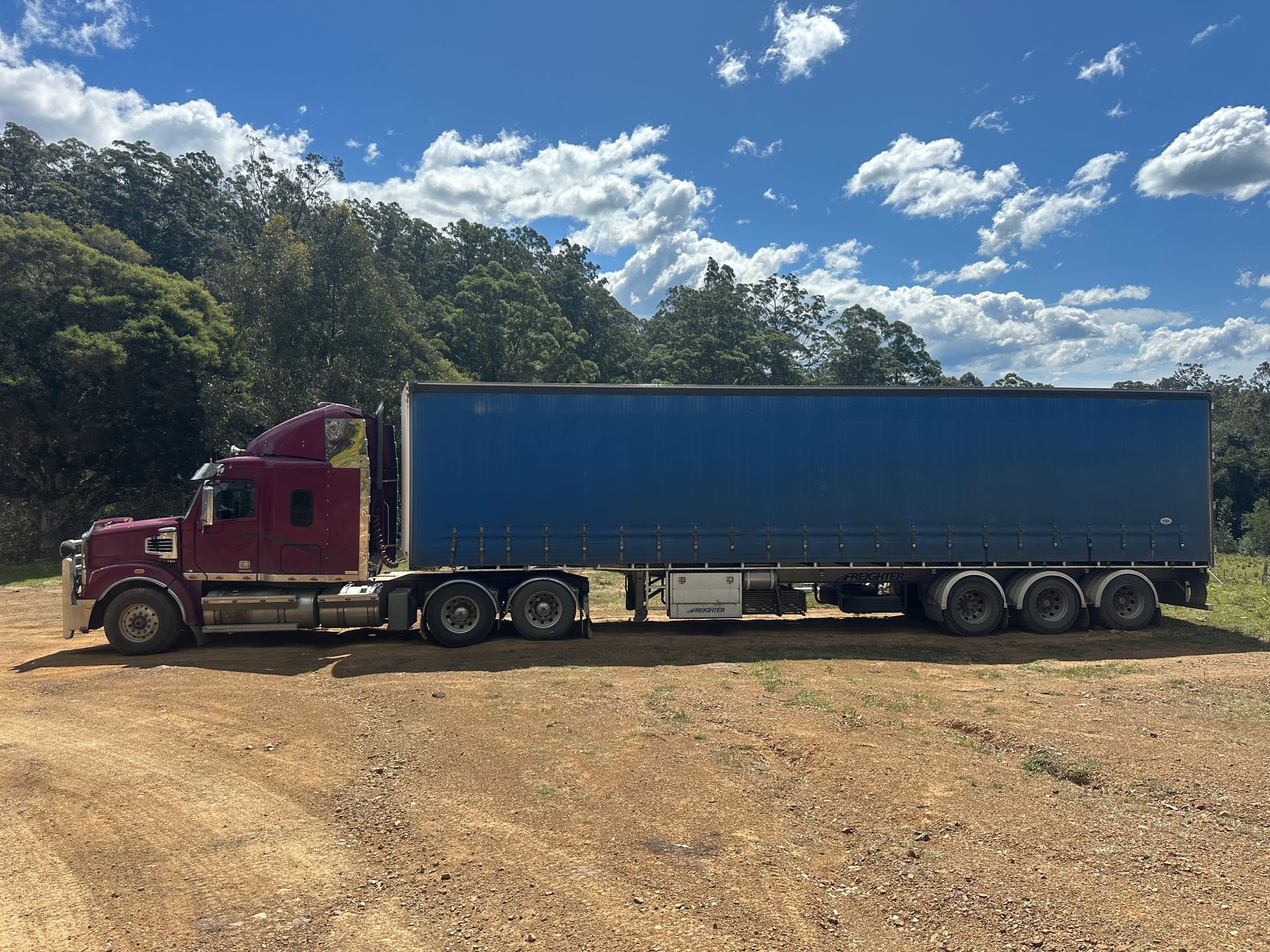Maroon semi-truck with blue trailer parked on dirt road, trees and blue sky background — D & M Freighters Water In Bulahdelah, NSW