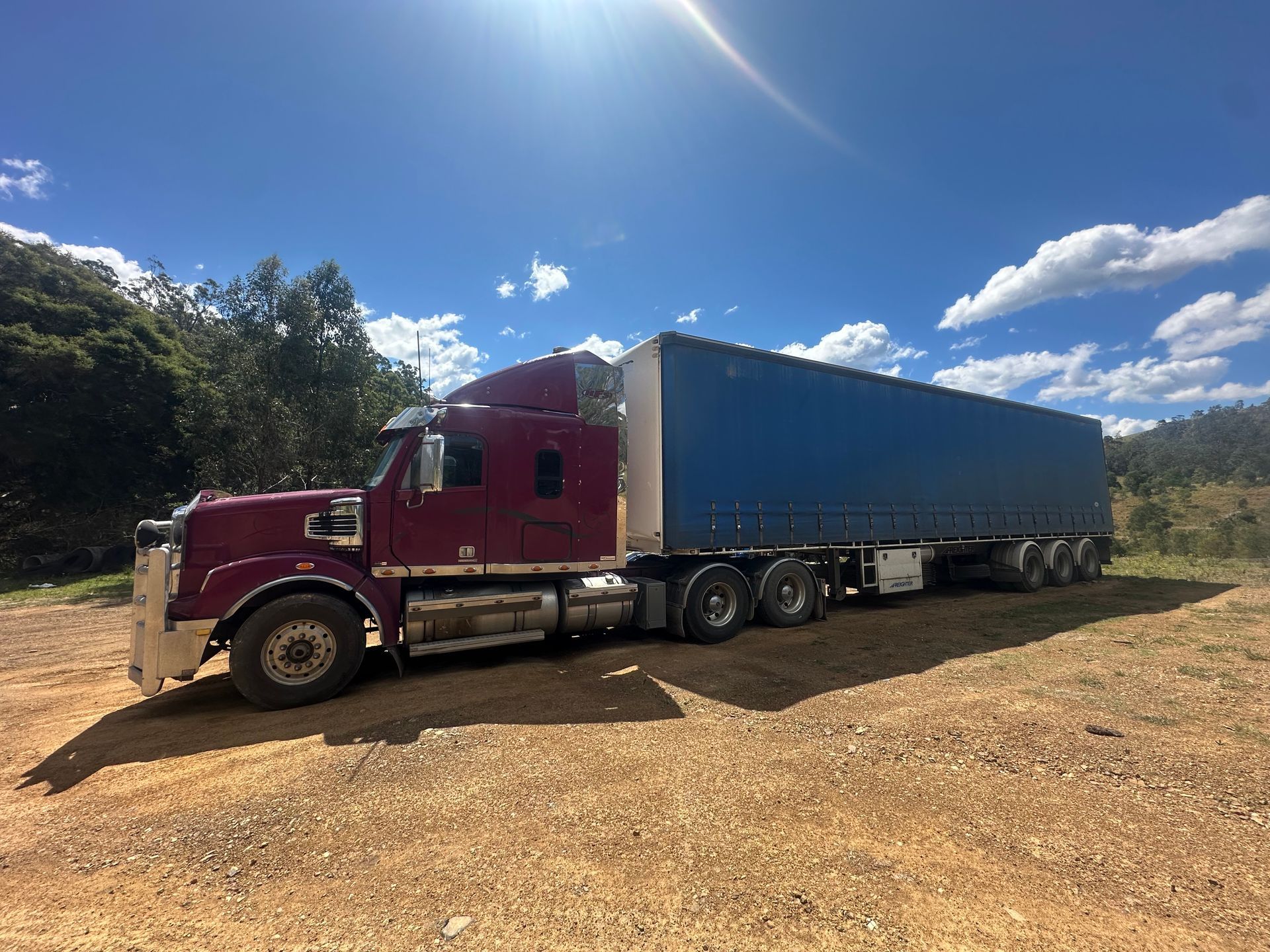 Burgundy semi-truck with a blue trailer parked on a dirt road in a sunny, natural setting — D & M Freighters Water In Forster, NSW