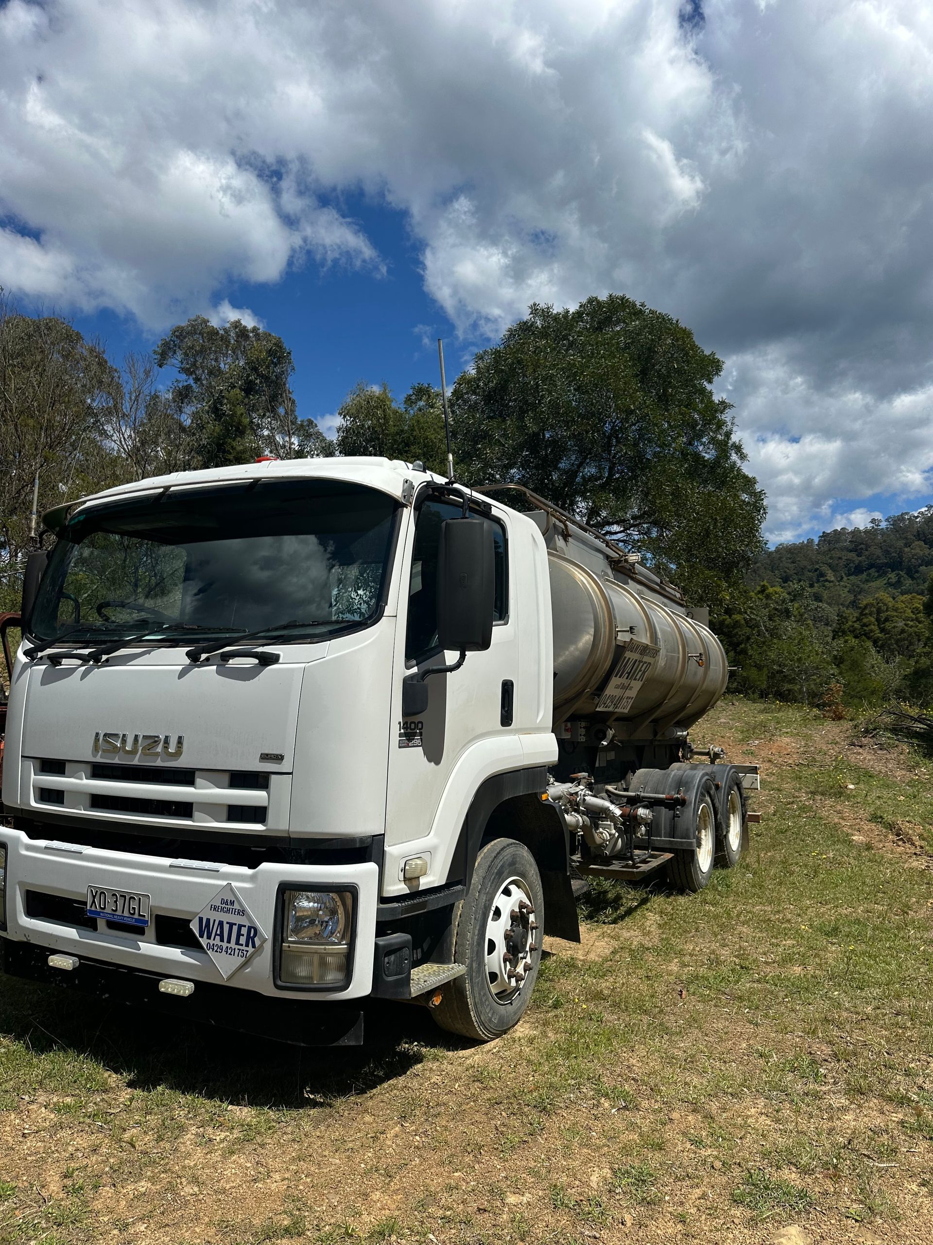 White Isuzu tanker truck parked on a grassy dirt road against a backdrop of trees and a cloudy sky — D & M Freighters Water In Bulahdelah, NSW