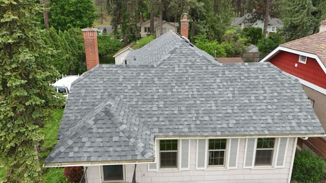Aerial view of a house with a gray shingled roof, brick chimneys, and surrounding tall green trees