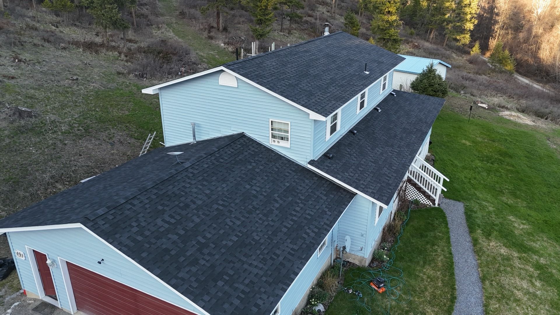 Aerial view of a light blue house with a dark roof beside a grassy yard and wooded hillside.