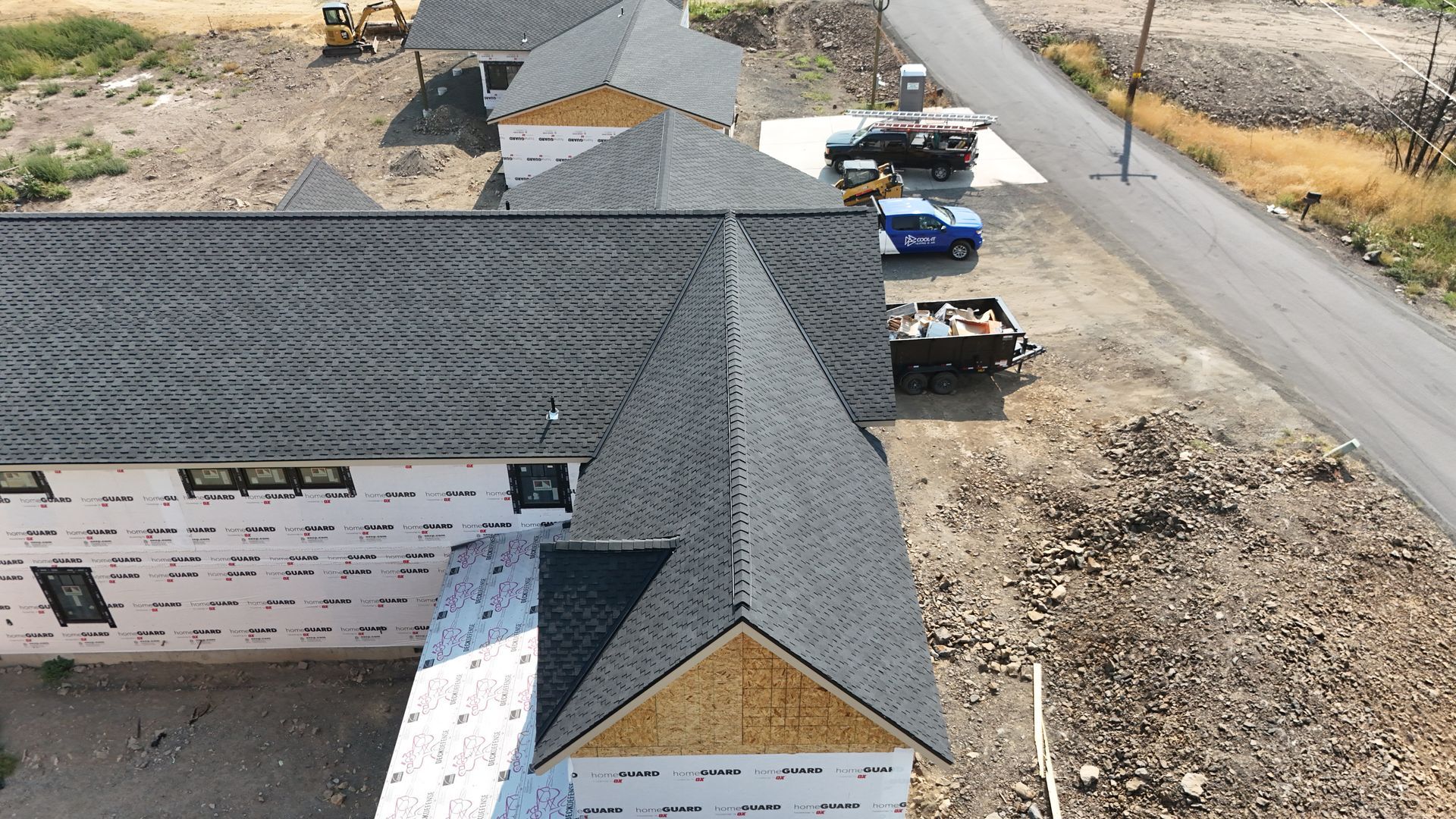 Aerial view of a house under construction with gray roof shingles, white siding, and trucks on a gravel lot.