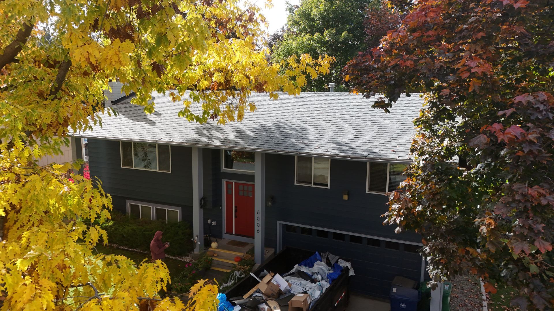 Two-story dark green house with red door, seen through yellow autumn trees and a driveway with bins.