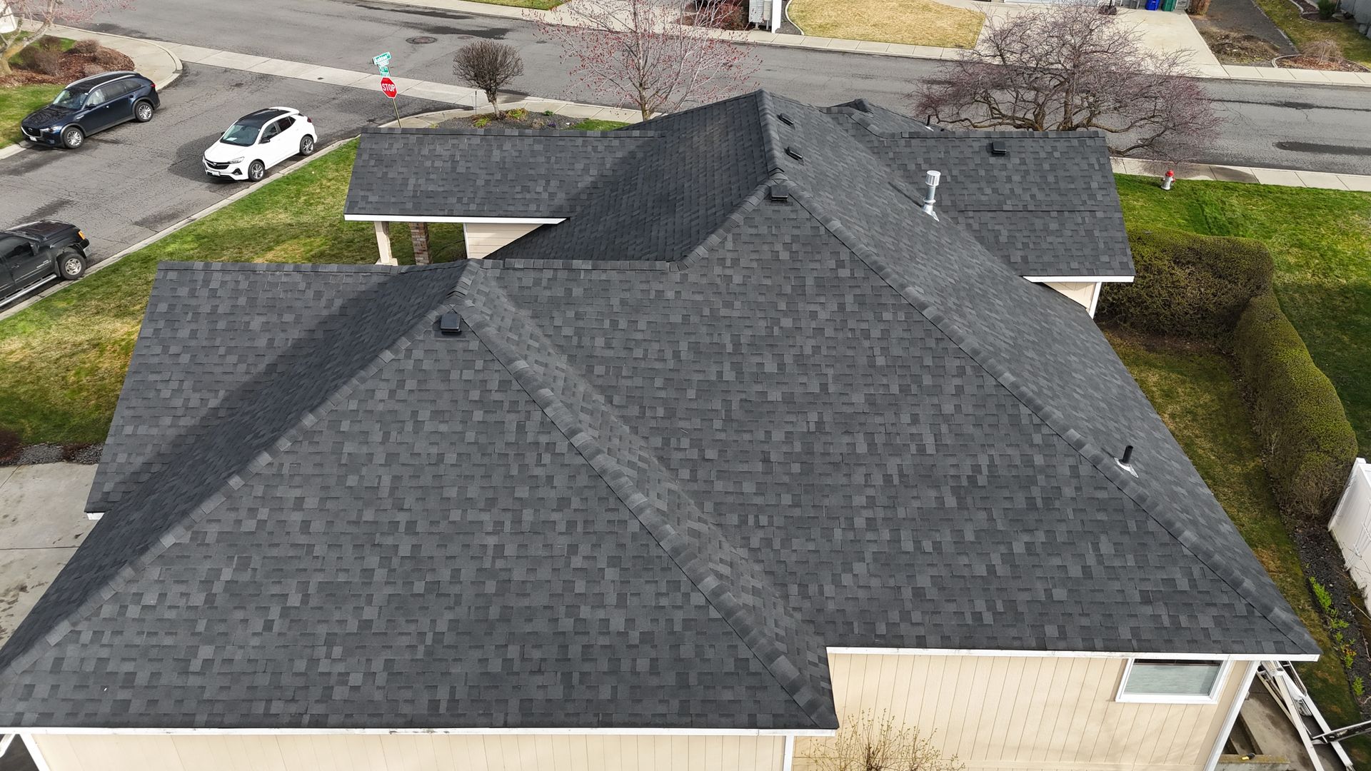 Aerial view of a house with a dark gray roof, driveway, parked cars, and nearby trees.