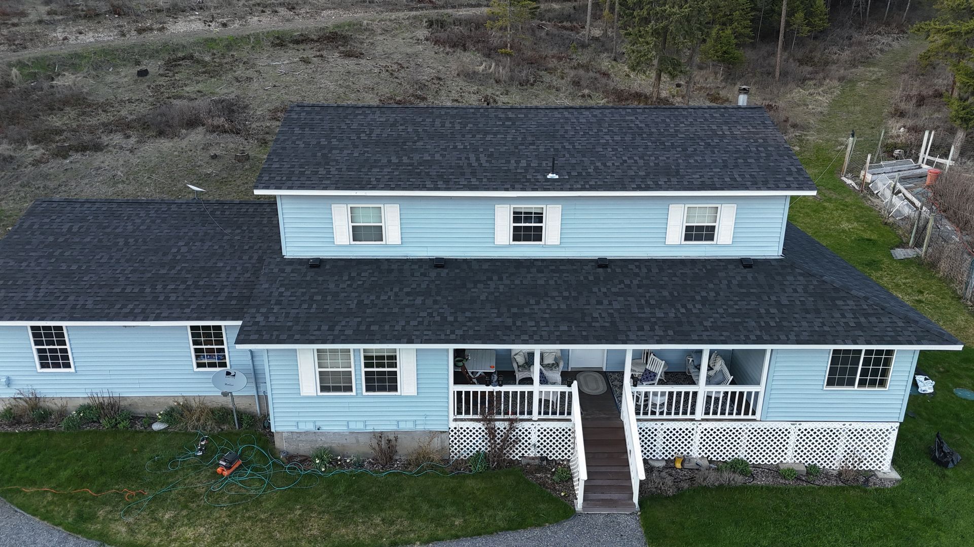 Aerial view of a light blue house with a dark roof and white porch on a wooded hillside