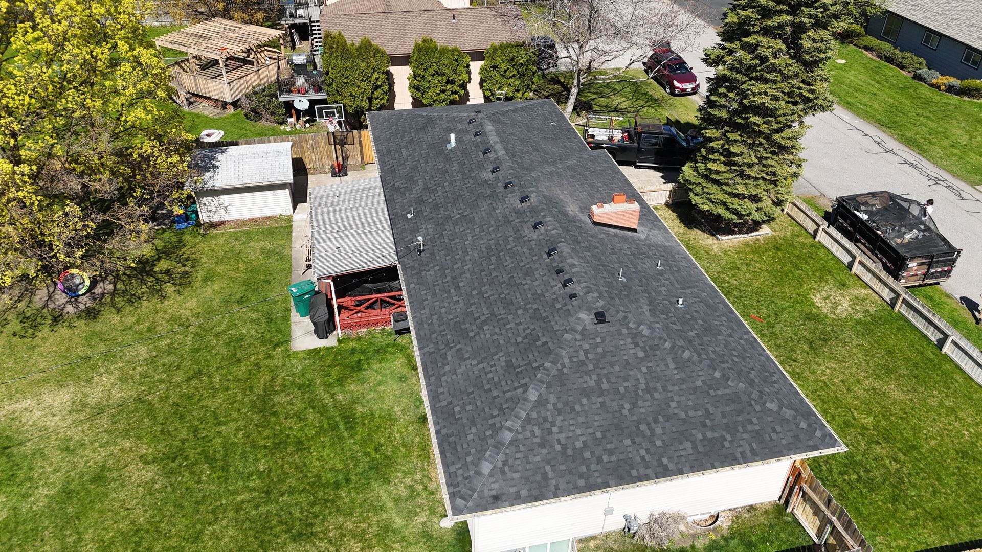 Aerial view of a house with a dark roof, green lawn, and backyard sheds beside a road and parked car.