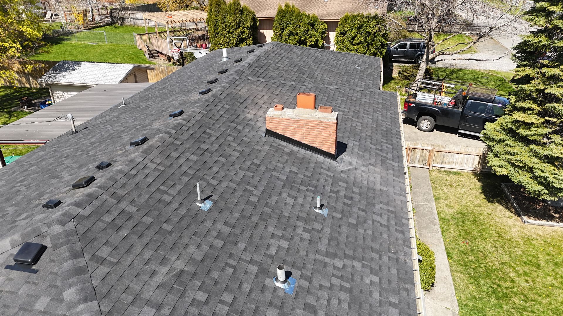 Aerial view of a house roof with black shingles, a brick chimney, and a driveway beside green landscaping.