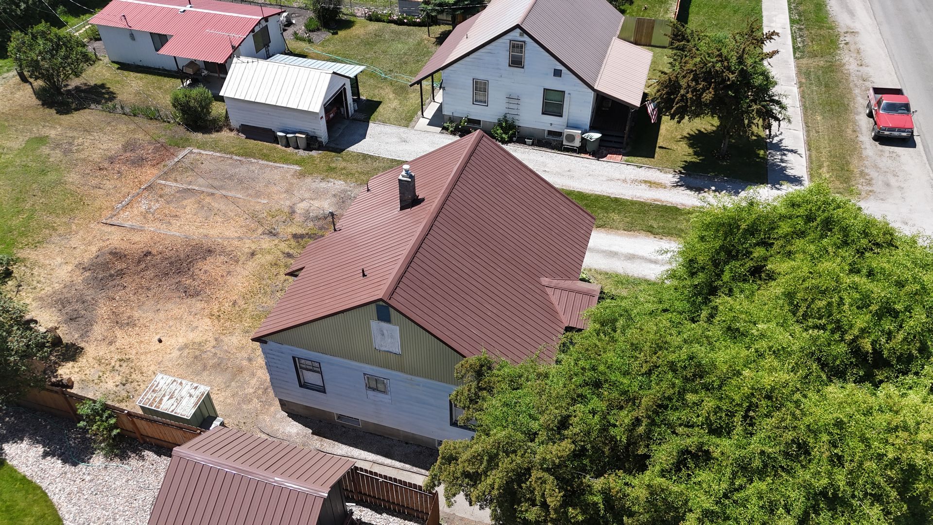Aerial view of a residential yard with several houses, red roofs, trees, and a parked red car on the road.
