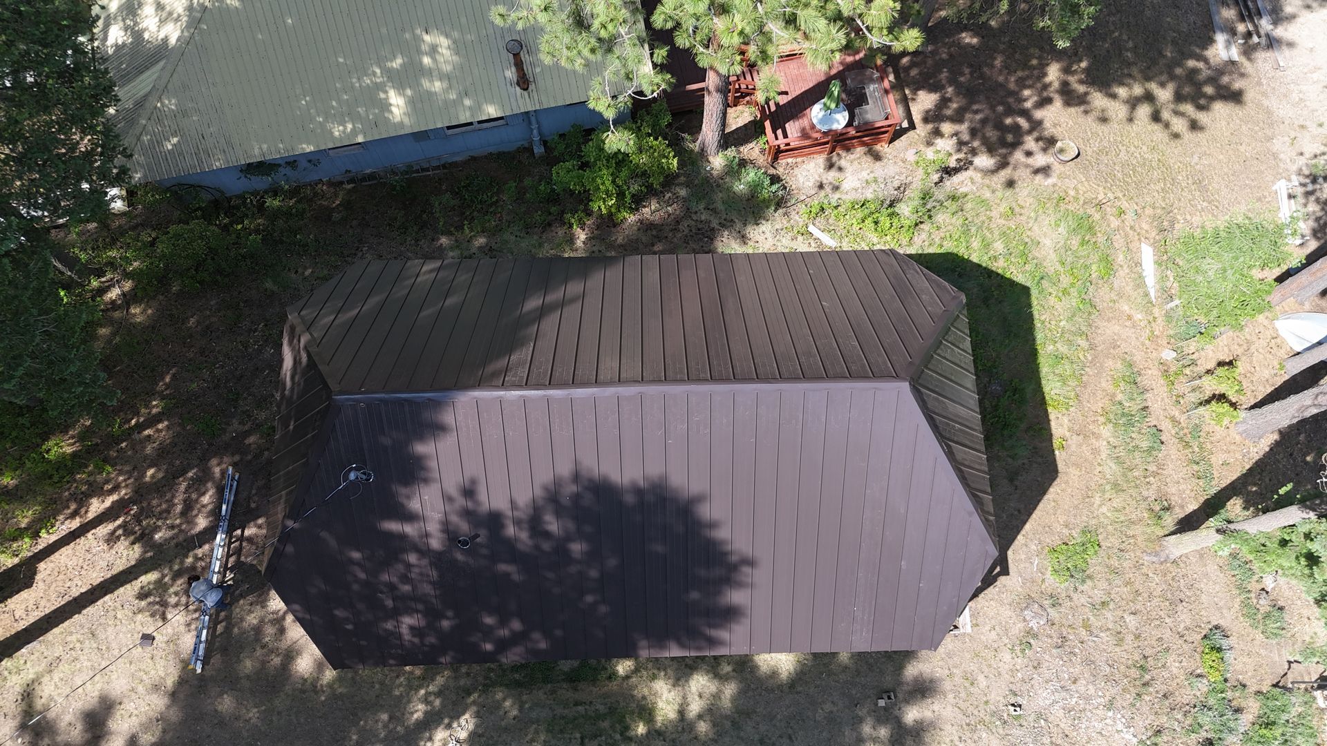 Aerial view of a small brown-roofed shed beside a tree and dirt ground, with shadows across the roof.