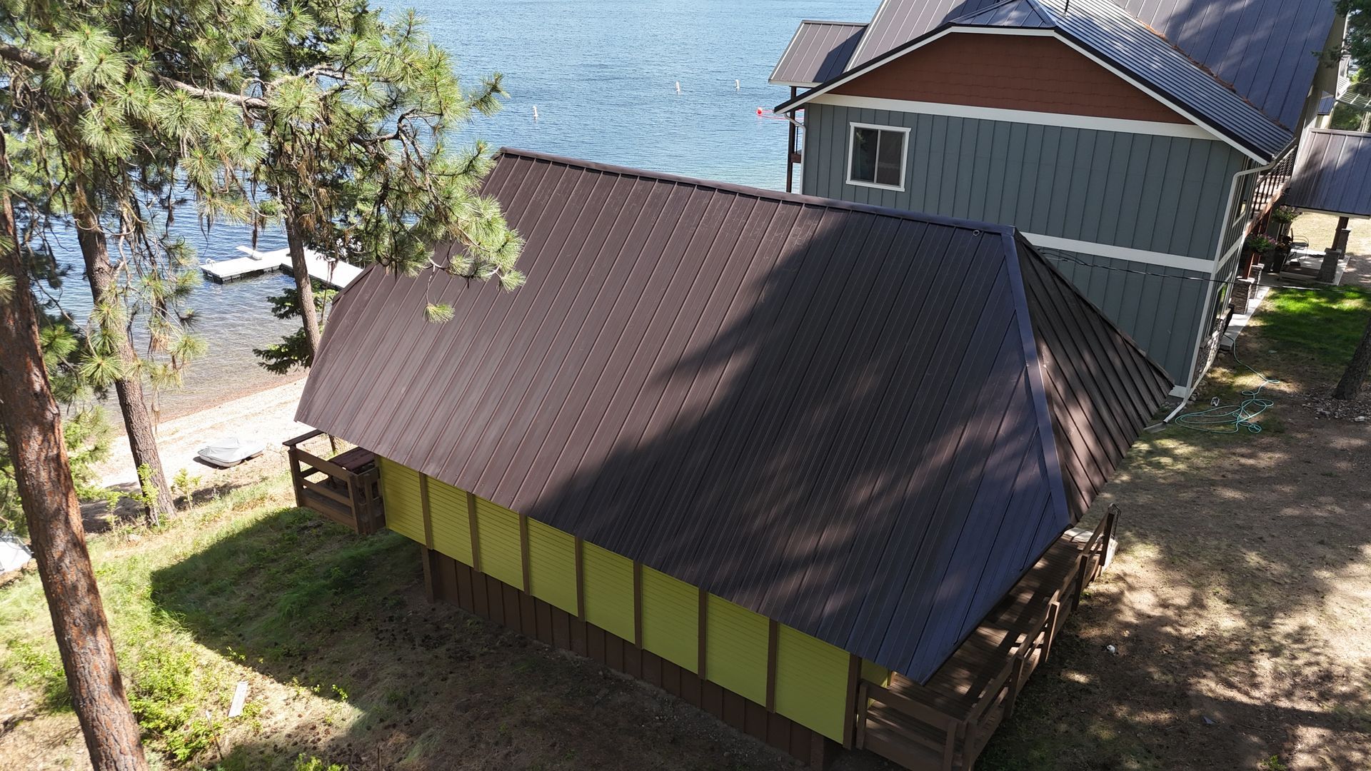 Aerial view of a brown-roofed cabin by the water, surrounded by trees and another house.