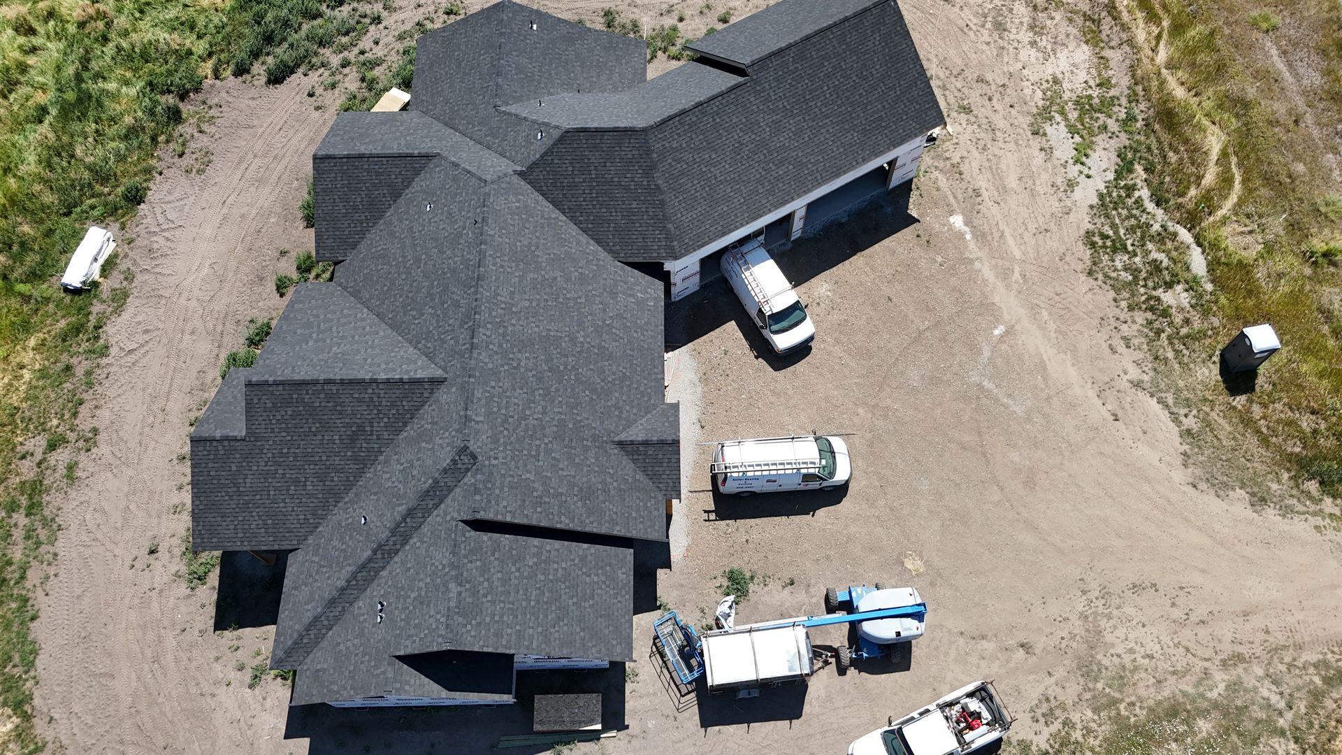Aerial view of a dark-roofed house with several vehicles parked on a dirt driveway and yard.