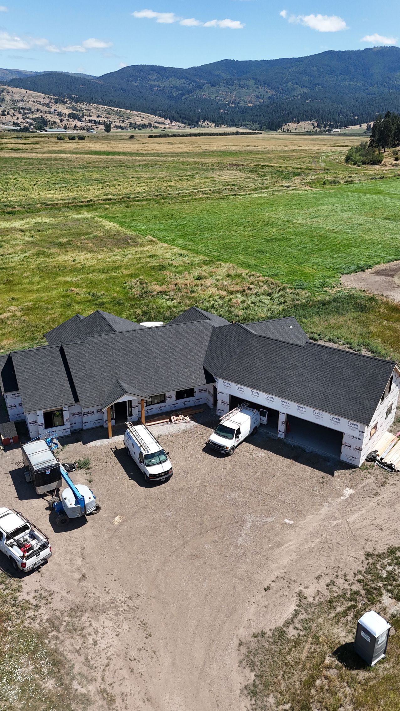 Aerial view of a rustic building with parked cars, set beside green fields and hills under a clear sky