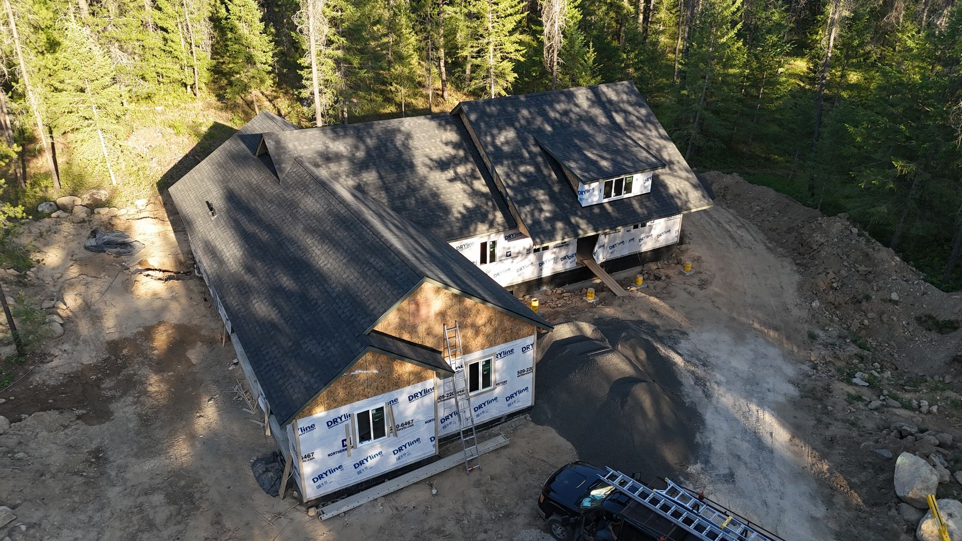Aerial view of two unfinished houses on a dirt lot surrounded by trees, with construction materials nearby.