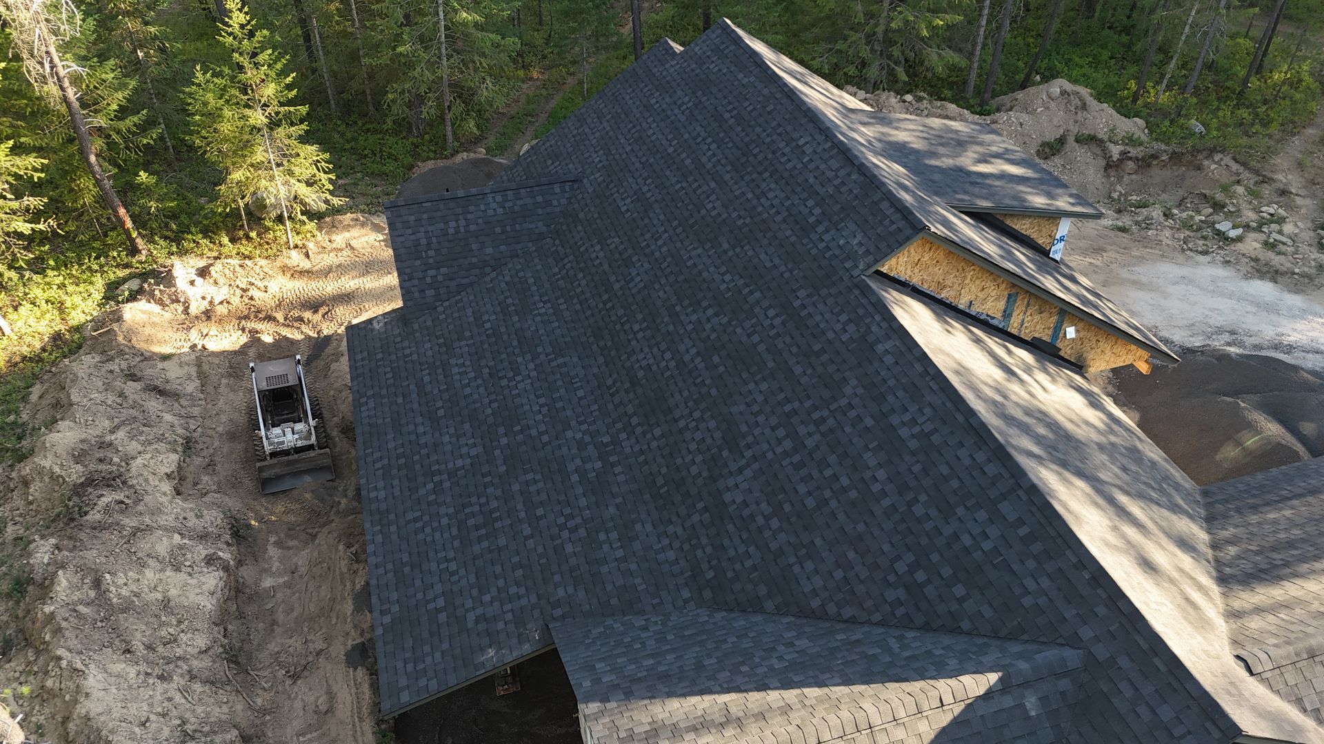 Aerial view of a dark shingled roof on a house under construction beside a wooded area