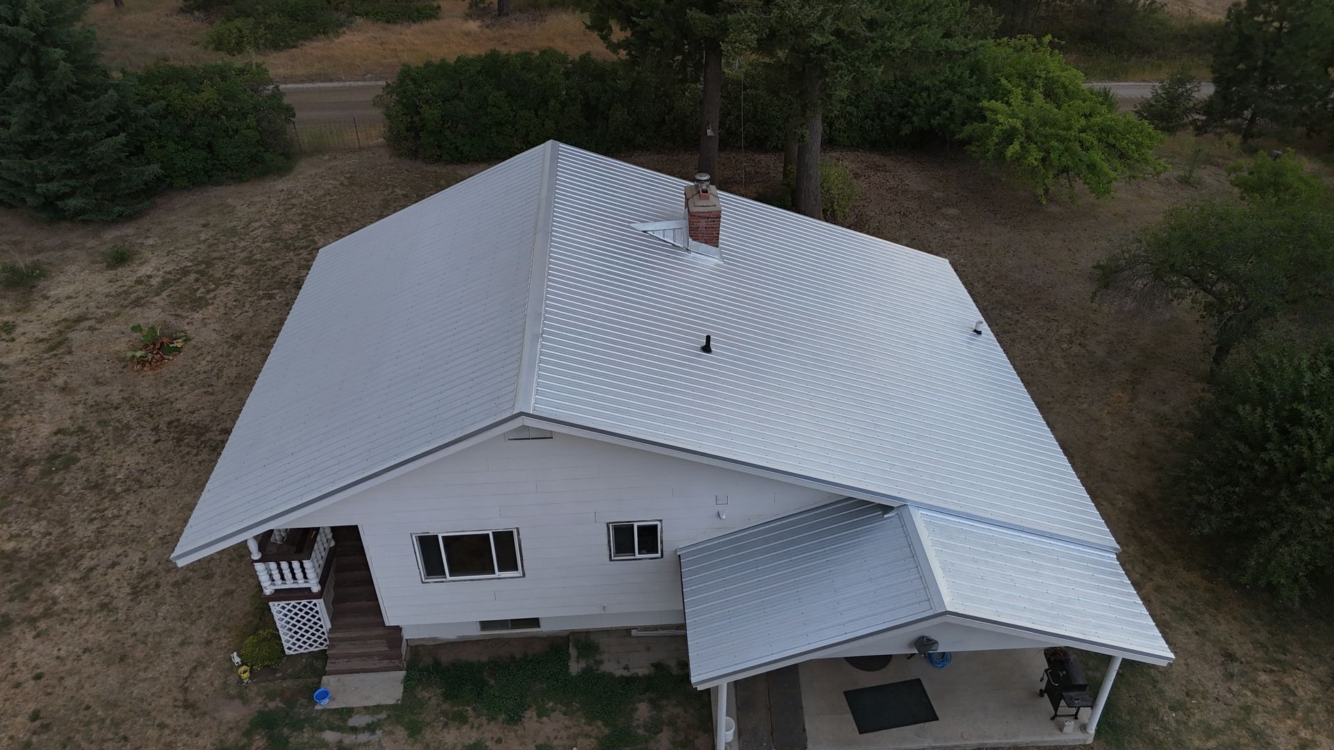 Aerial view of a white house with a metal roof surrounded by dry trees and yard.