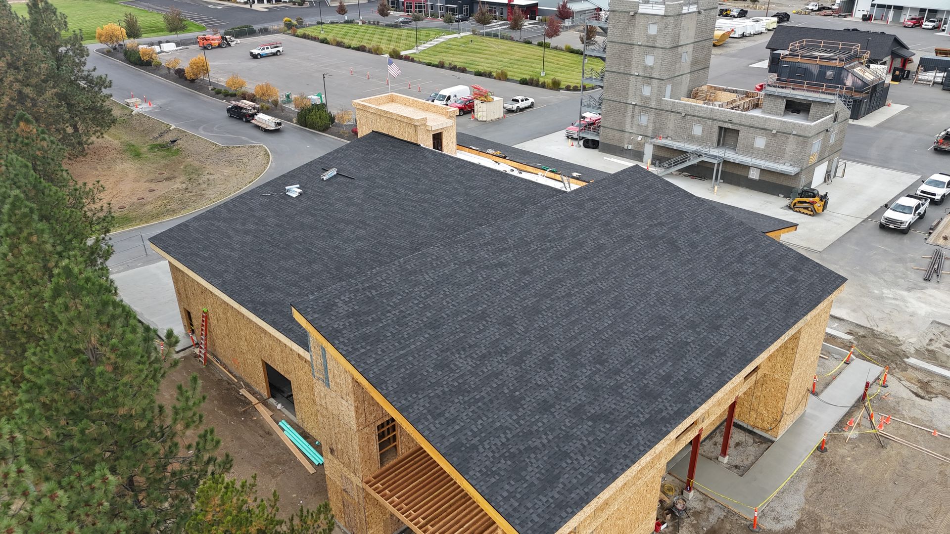 Aerial view of a building with a dark roof beside a parking lot and road near a stone church tower