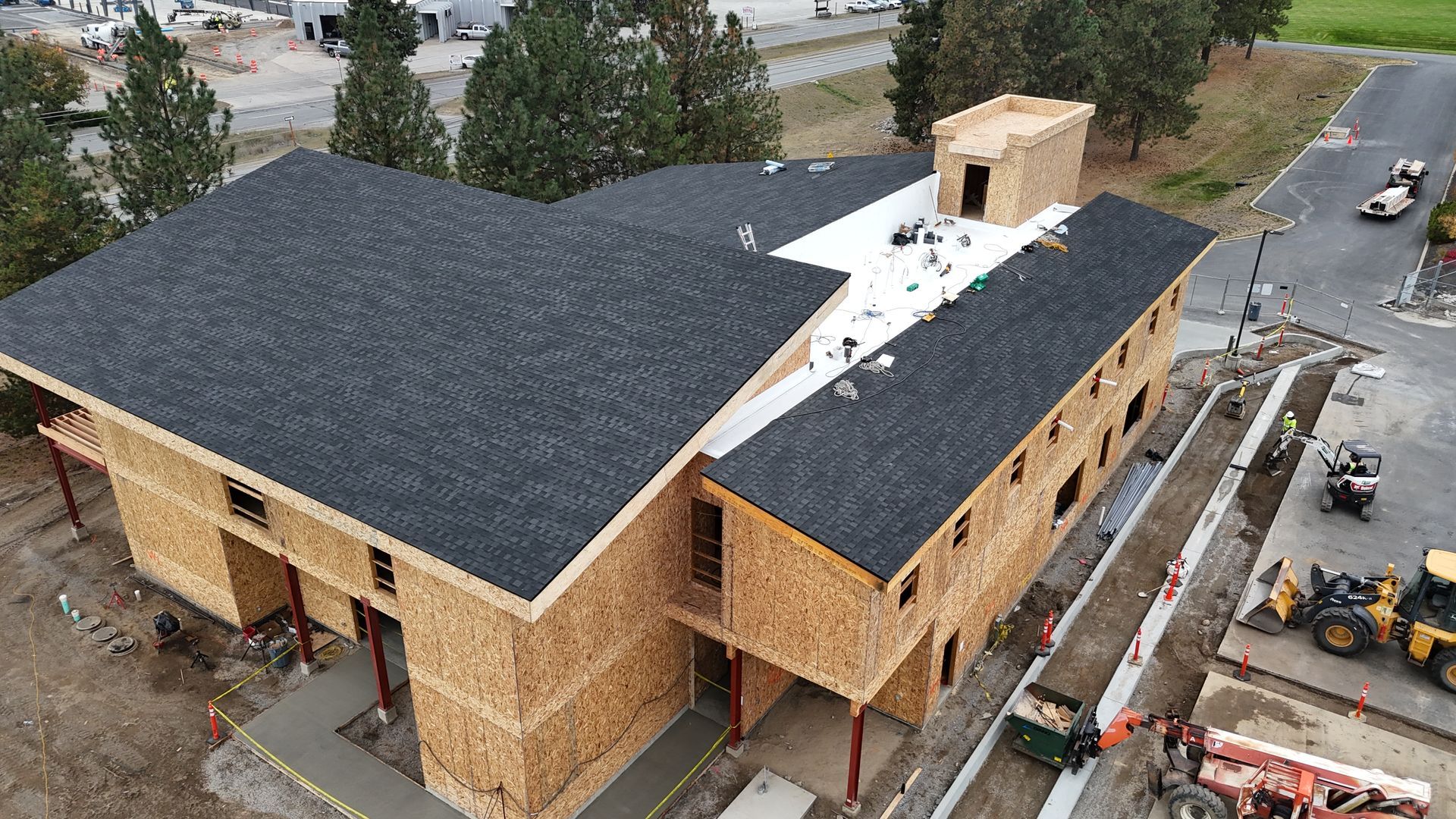 Aerial view of a wood-framed building under construction with dark roof shingles and surrounding work vehicles