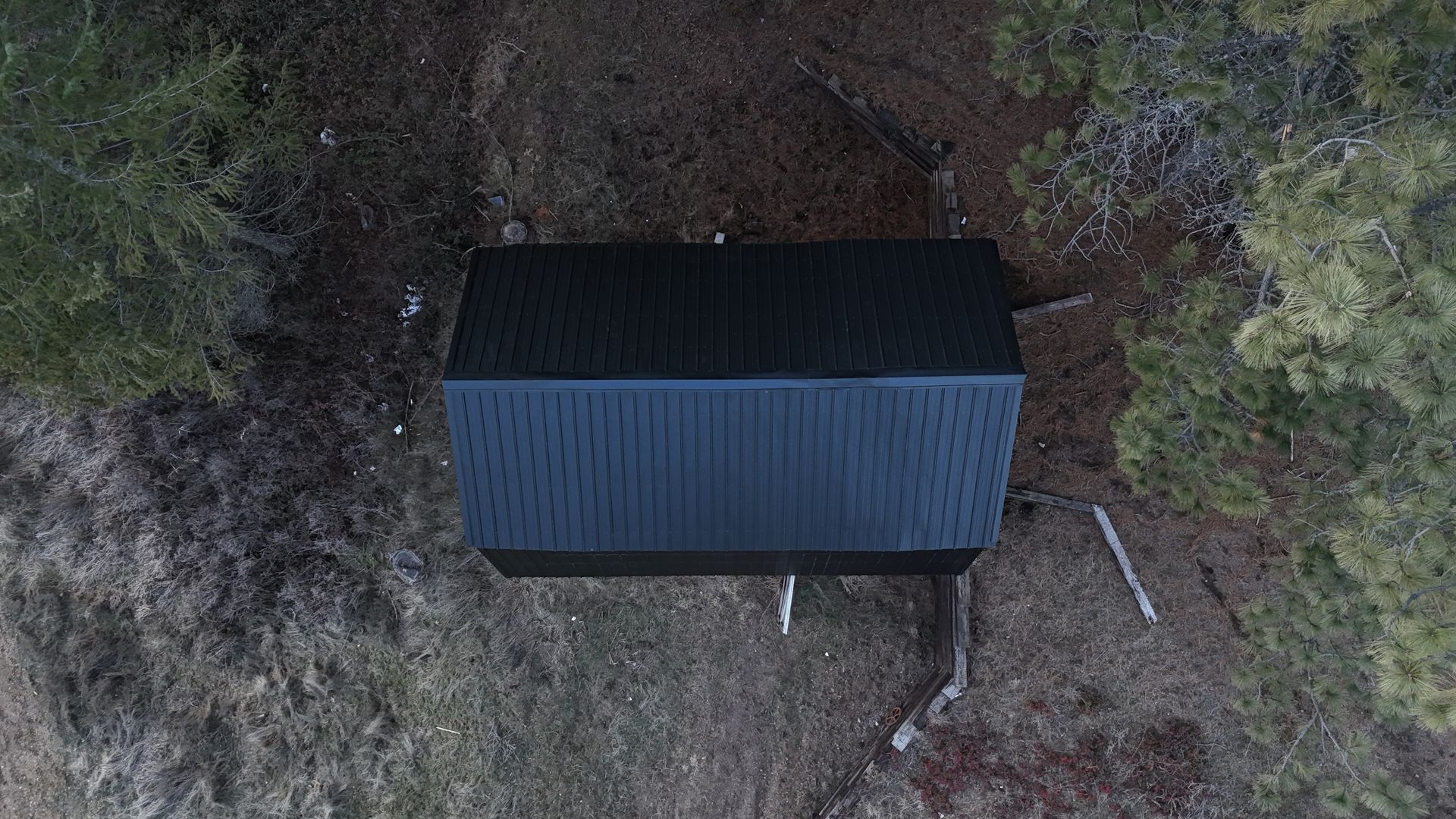 Top-down view of a small dark blue shed in a wooded clearing, surrounded by dirt and sparse trees.