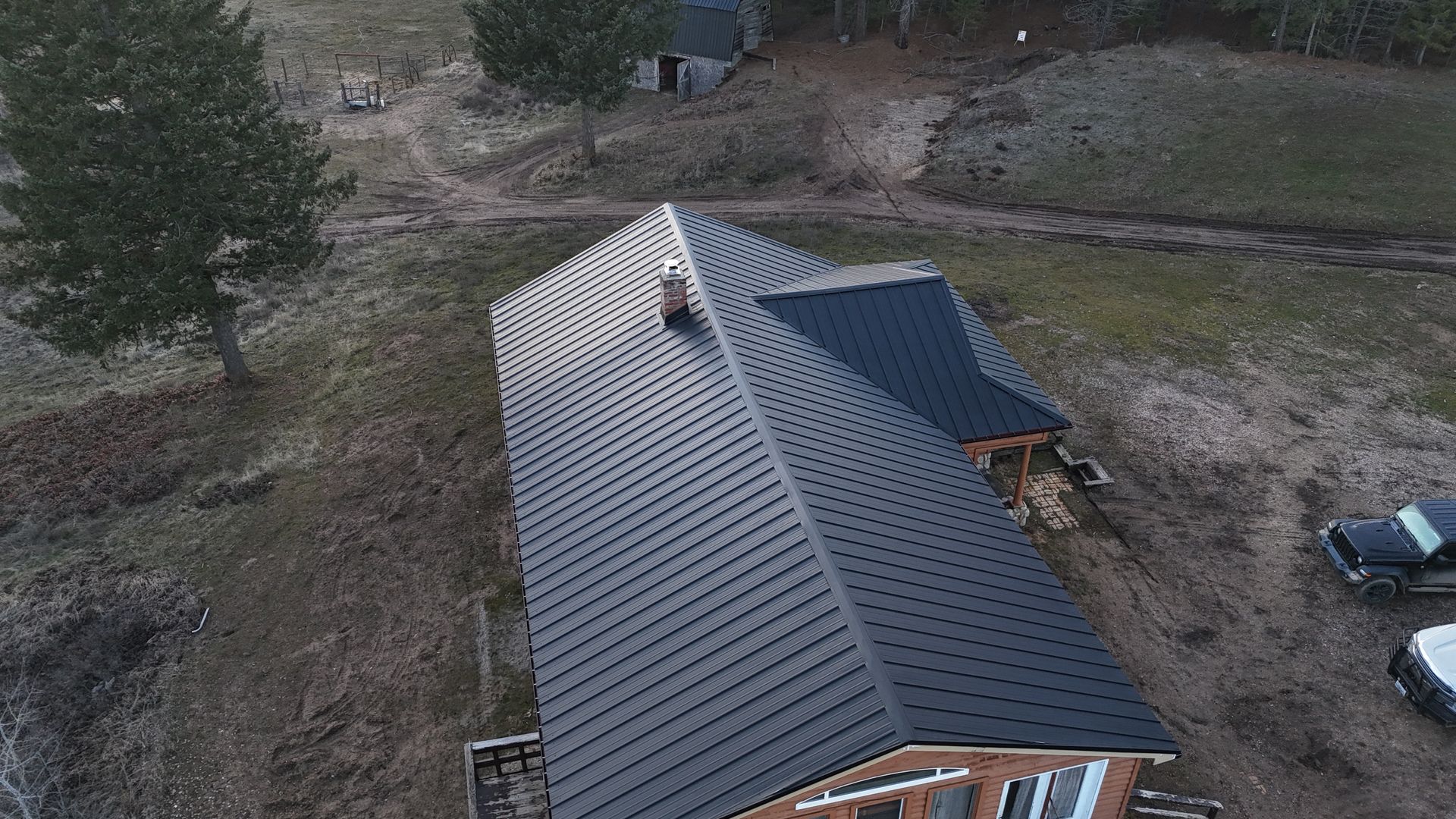 Aerial view of a metal-roofed house on a wooded property with vehicles nearby and a dirt yard