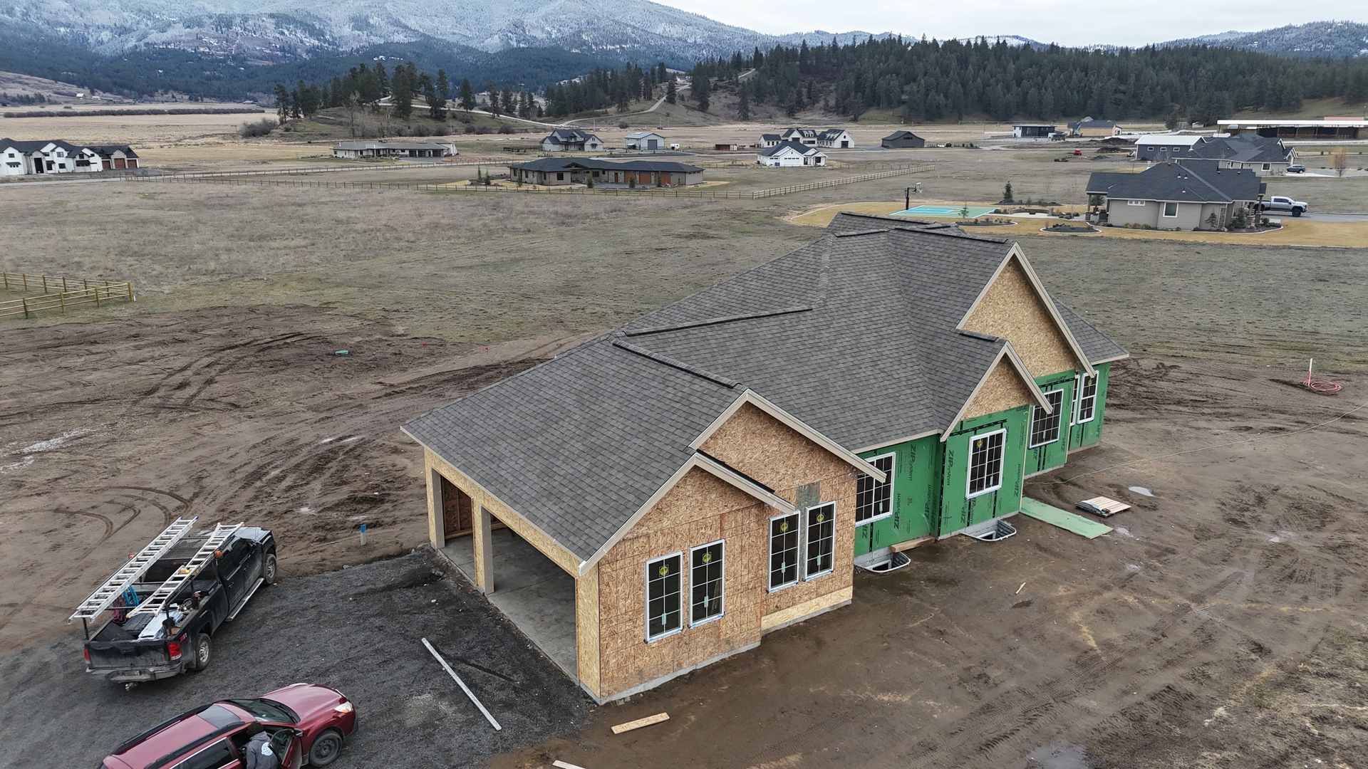 Partially built house with gray roof and green siding in a muddy rural lot, mountains in the background