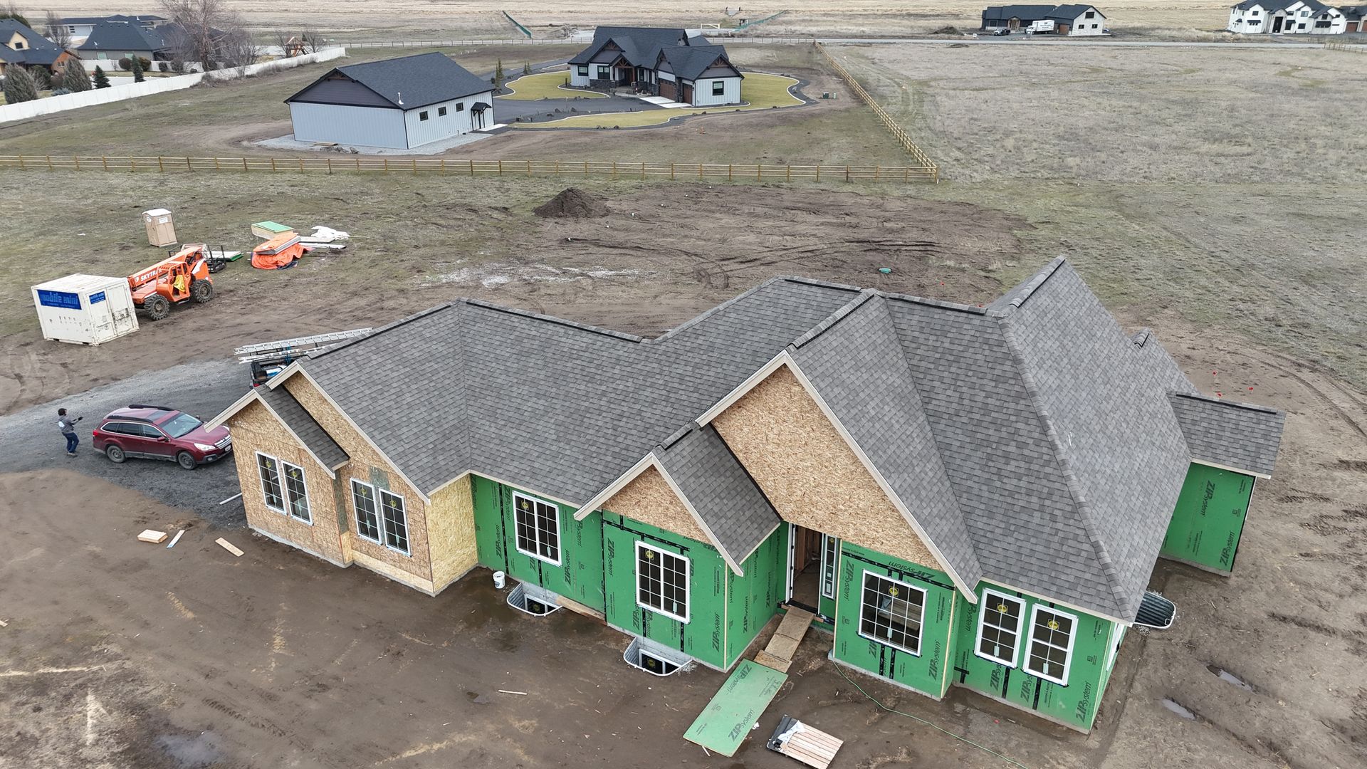 Aerial view of a green-and-tan house under construction on a dirt lot with scattered equipment nearby