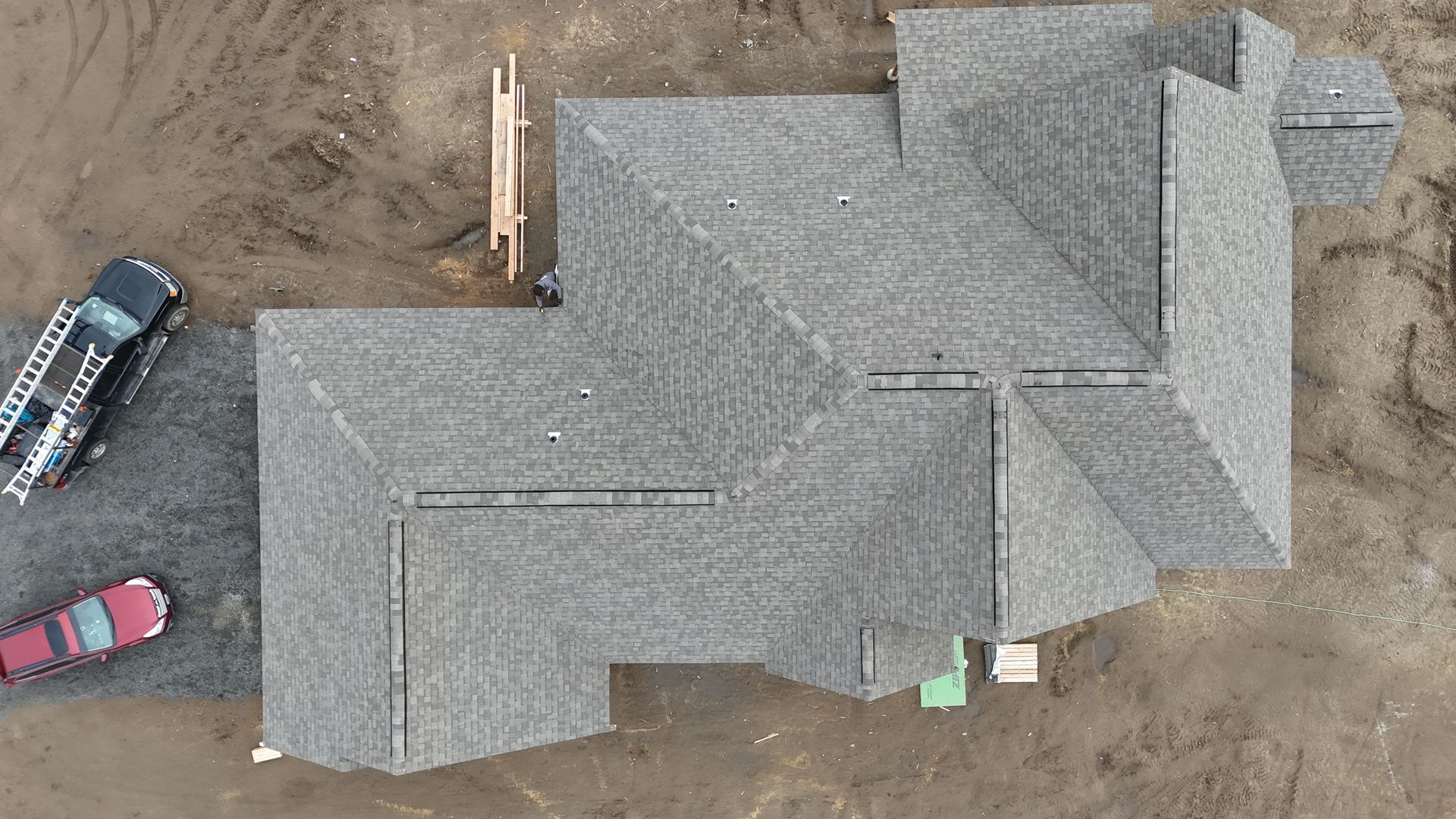 Aerial view of a gray shingled house roof with parked vehicles beside it on dirt ground