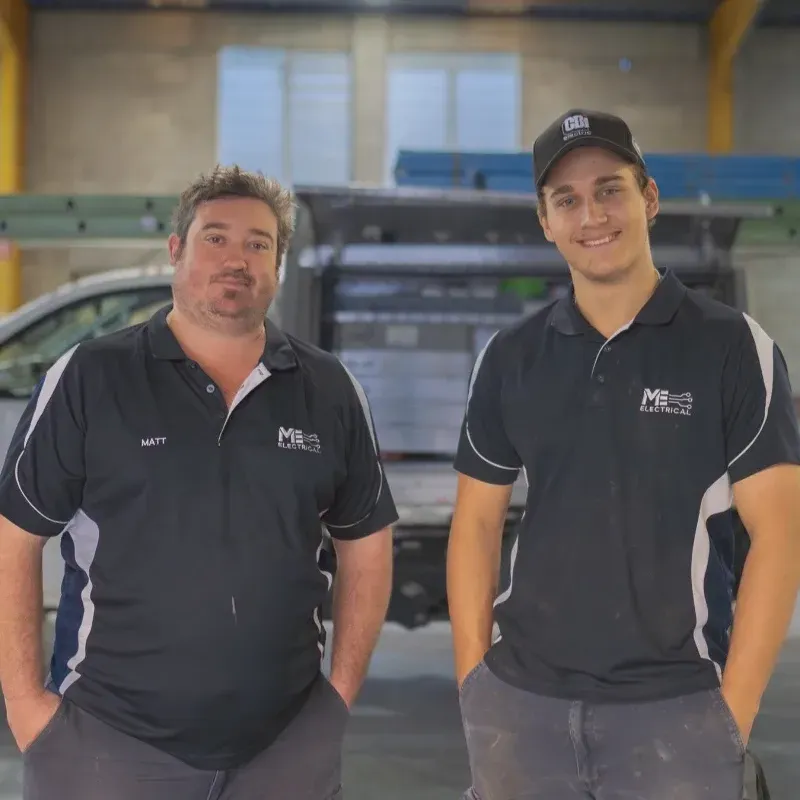 Two electricians are standing in front of a work ute in a warehouse.