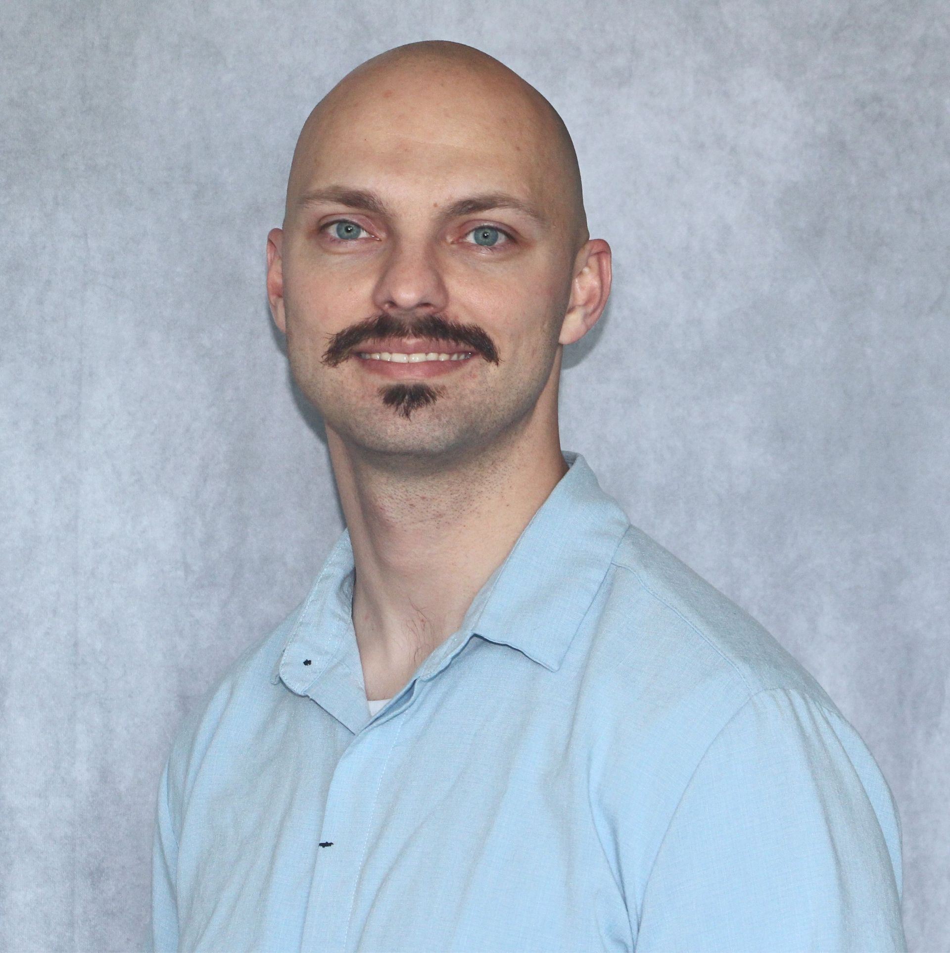 Portrait of a bald man with a mustache in a light blue shirt against a gray backdrop