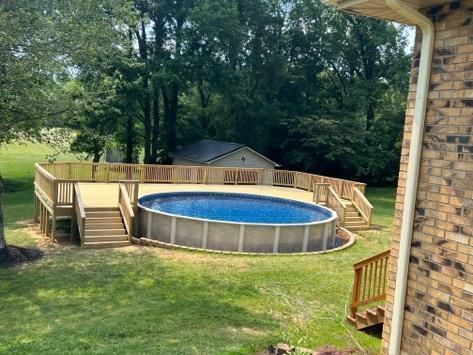 Above-ground pool with wooden deck in backyard. Green grass, trees, and a brick house are visible.
