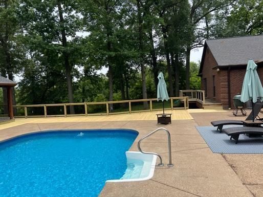 Pool with blue water, patio, and low wooden fence in a backyard setting, trees in the background.
