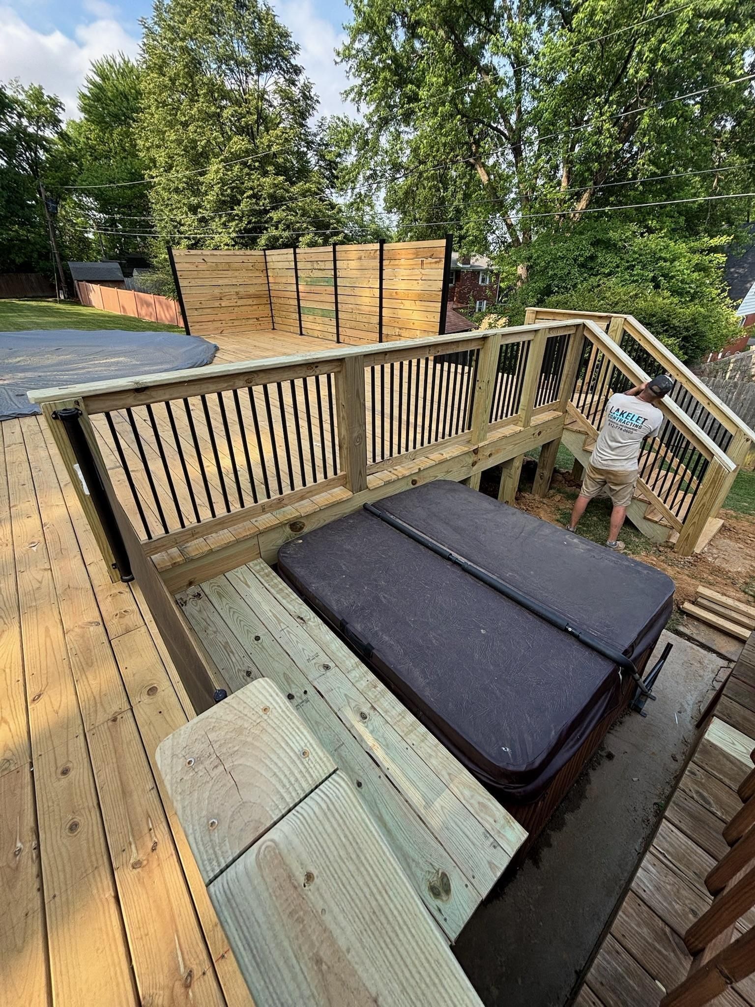Hot tub on a wooden deck with railings, steps, and a privacy fence. A person works near the stairs.