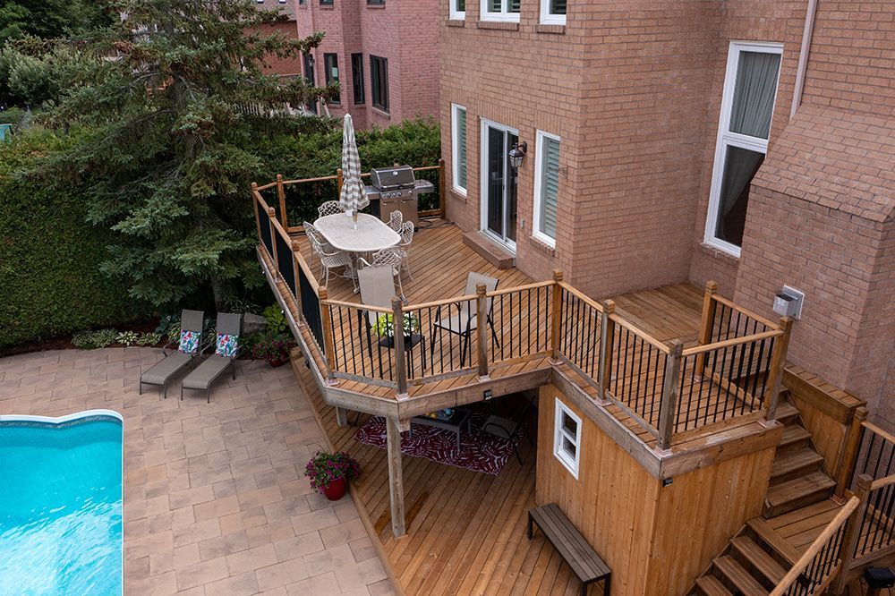 Wooden deck with outdoor dining set, grill, and lounge chairs, overlooking a pool and brick house.
