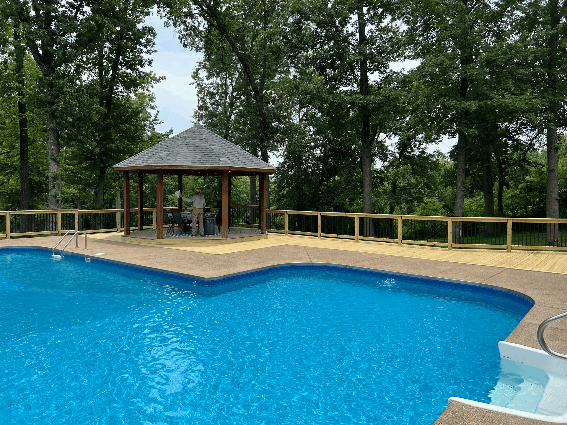 Swimming pool with a gazebo, surrounded by a wooden fence and trees. Blue water, beige decking, and a cloudy sky.