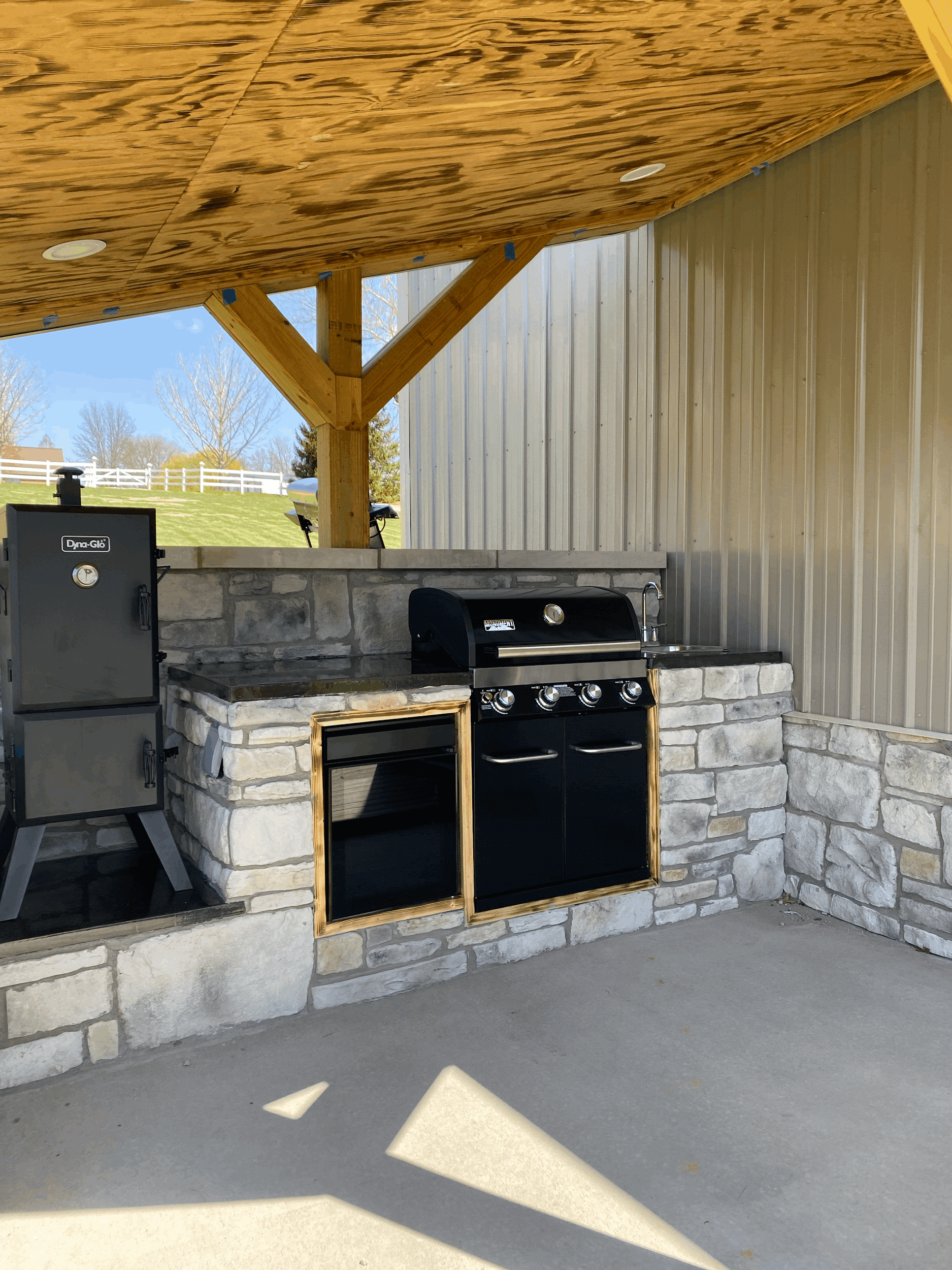 Outdoor kitchen with smoker, grill, and stone surround under a wooden roof.