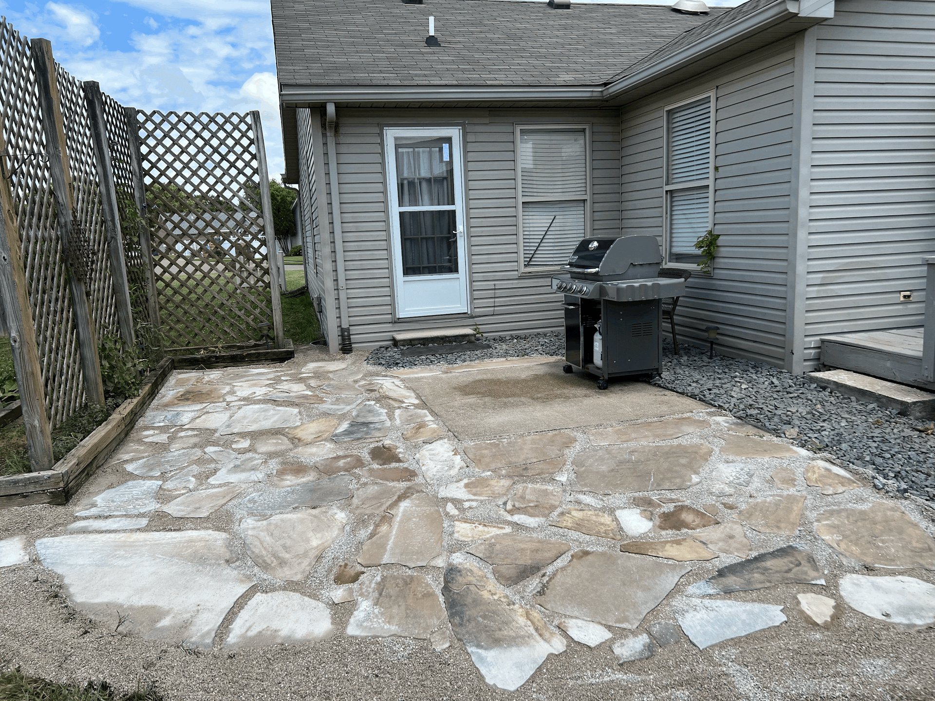 Stone patio with grill next to a house. Grey walls, white door and window, wooden fence.