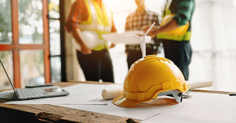 Yellow hard hat on a table with blueprints, laptop, and three construction workers in the background.