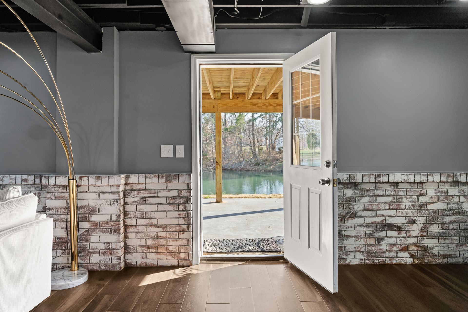 Basement interior with open white door leading to an outdoor deck over water. Gray walls, exposed beams, and brick.
