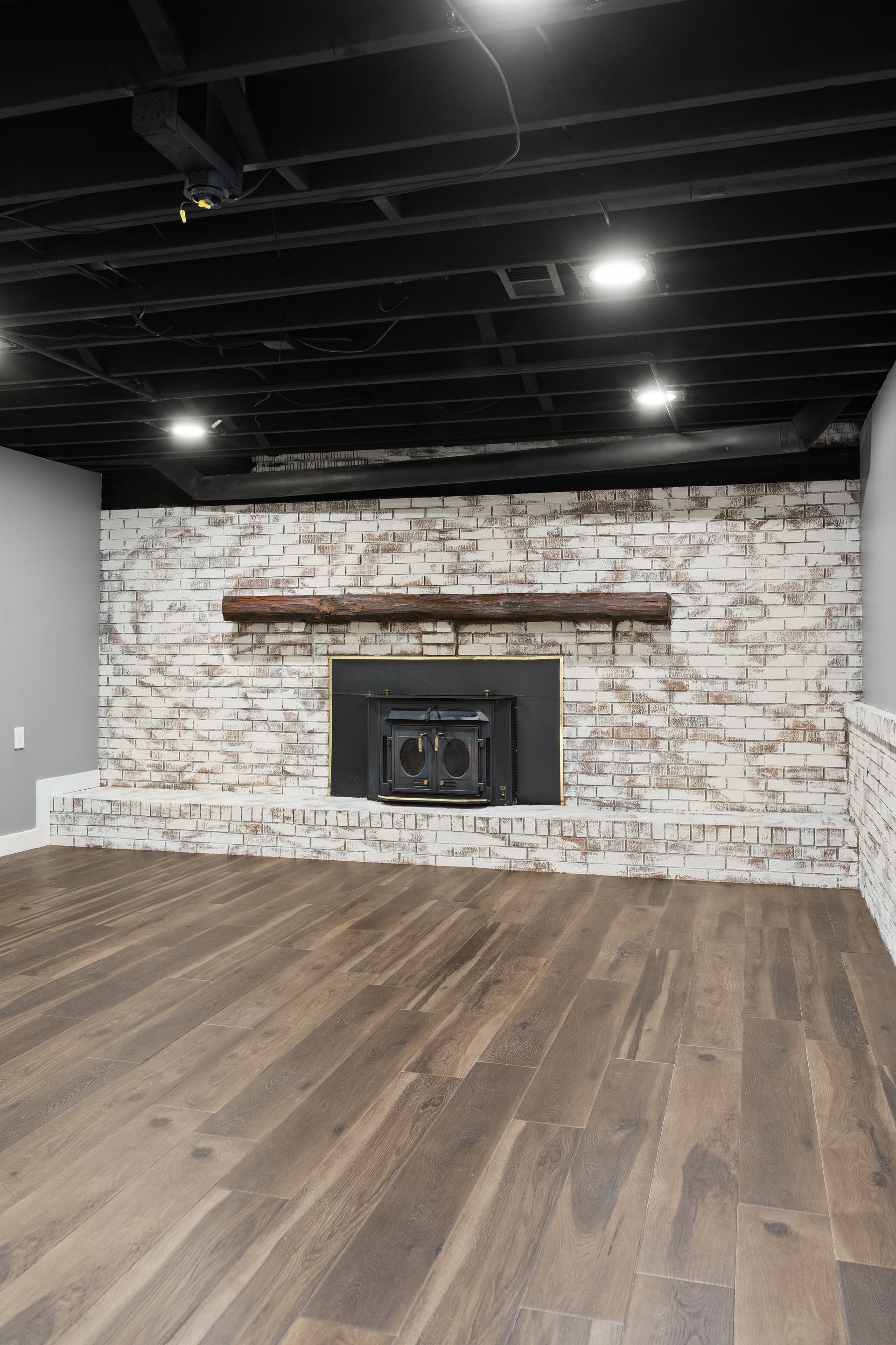 Fireplace with faux brick, dark mantel, in a basement with wood floor and black ceiling.