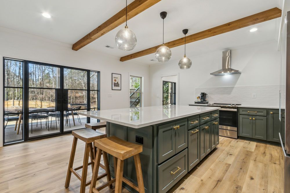 Spacious kitchen with a green island, wooden beams, and large windows overlooking a patio and trees.
