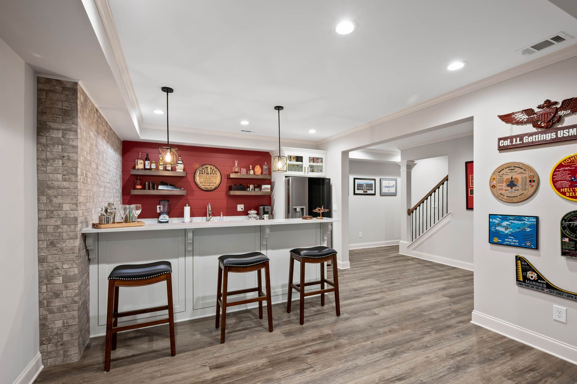 A basement bar with seating, red accent wall, stone-like side wall, and military decor. Flooring is wood-like. Stairs lead to another area.